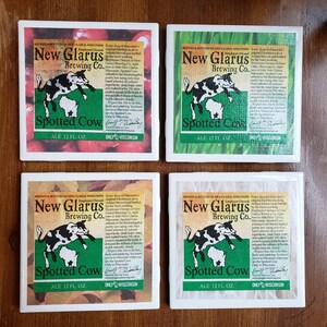 May include: Four white square coasters with a green, white and red design. Each coaster features a cartoon spotted cow jumping over the state of Wisconsin. The text "New Glarus Brewing Co." and "Spotted Cow Ale 12 fl. oz." is printed on each coaster. The text "Only in Wisconsin" is printed on the bottom of each coaster.