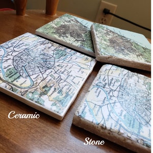 May include: Three square coasters featuring vintage map prints of Paris and its surrounding suburbs. One coaster is smooth white ceramic, while the other two have a textured, porous stone edge. The maps display street grids, rivers, and city names.