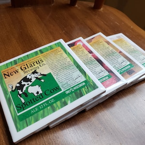 May include: A stack of four white square coasters with green and white beer labels. The top coaster features a spotted cow illustration and the text "New Glarus Brewing Co. Spotted Cow Ale 12 fl. oz. Only Wisconsin".