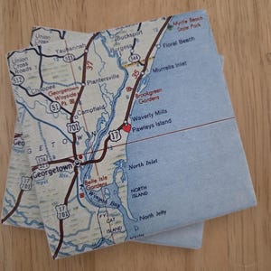 May include: Two square coasters featuring a printed map of the South Carolina coast, including Georgetown, Pawleys Island, and Murrells Inlet. The map displays roads, water bodies, and a red heart icon marking Pawleys Island on a light background.