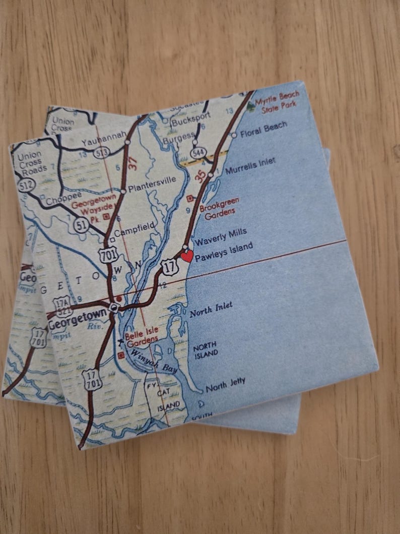 May include: Two square coasters featuring a printed map of the South Carolina coast, including Georgetown, Pawleys Island, and Murrells Inlet. The map displays roads, water bodies, and a red heart icon marking Pawleys Island on a light background.