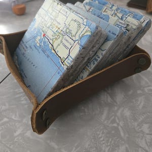 May include: A set of square stone coasters featuring printed map details of Michigan, including Mackinac and Goetzville, held within a brown leather tray with metal snap fasteners at the corners. The coasters have a textured, light grey stone edge.
