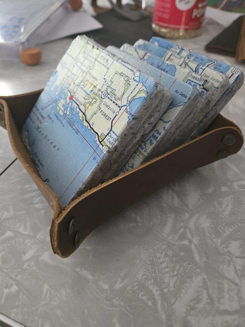 May include: A set of square stone coasters featuring printed map details of Michigan, including Mackinac and Goetzville, held within a brown leather tray with metal snap fasteners at the corners. The coasters have a textured, light grey stone edge.