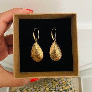 Peut inclure: Une paire de boucles d'oreilles pendantes dorées avec un design texturé et ondulé. Les boucles d'oreilles sont présentées dans une boîte brune avec un intérieur noir.