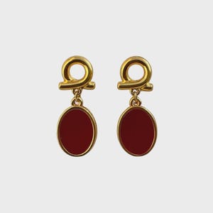 Peut inclure: Une paire de boucles d'oreilles dorées. Chaque boucle d'oreille présente une boucle dorée en haut, reliée à une pierre rouge foncé ovale, encadrée d'or. Les boucles d'oreilles sont présentées sur un fond clair et uni.