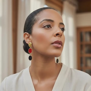 Peut inclure: Boucles d'oreilles pendantes dorées avec un disque supérieur texturé, un ovale rouge et un ovale noir. Les boucles d'oreilles sont portées par une personne aux cheveux foncés et un haut blanc. Bijou audacieux.