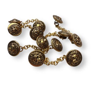 May include: Gold-toned metal buttons with lion head details, connected by delicate chains. The buttons are round with a raised lion face design. The chains and buttons have a vintage aesthetic, suitable for clothing or craft projects.