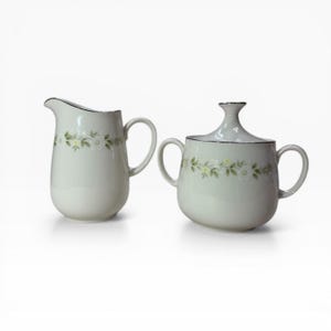 May include: A white porcelain creamer and covered sugar bowl set. Both pieces feature a floral design with white flowers and green leaves. The rims are trimmed with a thin black line. The creamer has a handle and spout.
