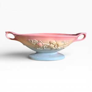 May include: A pastel-colored ceramic centerpiece bowl with a floral design. The bowl features a pink and peach rim, a light blue base, and two loop handles. The floral relief includes yellow and white flowers.
