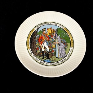 May include: White ceramic plate with a colourful illustration from "The Tinder Box" by Hans Christian Andersen. The plate features a soldier and a witch in a garden scene, with text around the border. The plate is from 1972.