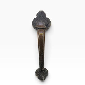 May include: A dark bronze-toned metal door handle featuring a curved grip and two decorative mounting plates. The plates have a pointed, scalloped shape with a subtle grid pattern etched into the surface, showing signs of aged patina.