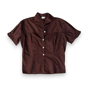May include: A short-sleeved, dark brown button-down shirt. The shirt features a classic collar, pleats down the front, and white buttons. The fabric appears to be a smooth, solid color, suitable for a vintage or retro aesthetic.