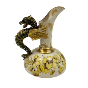 May include: A ceramic pitcher featuring a cream-colored base with a yellow floral pattern and a crackled glaze finish. The handle is shaped like a dark green and gold dragon with wings. The neck of the pitcher has gold accents.