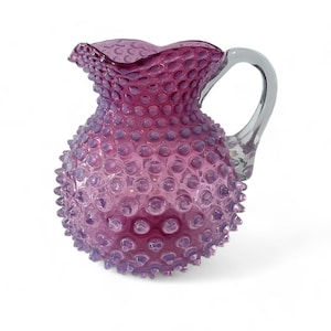 May include: A purple hobnail glass pitcher with a clear handle. The pitcher has a ruffled rim and a textured surface covered in small, rounded bumps. The handle is smooth and curved, attached to the side of the pitcher.