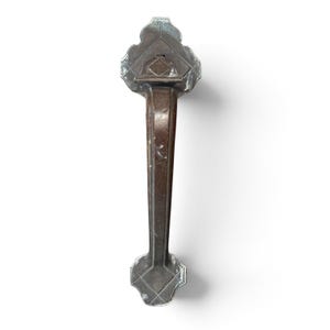 May include: A dark brown metal door handle featuring a vertical grip and two decorative plates at the top and bottom. Each plate has a geometric, multi-faceted shape with a central diamond-like engraving and worn, silver-toned edges.