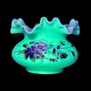 May include: A green glass vase with a ruffled rim and a bulbous body. The vase is decorated with hand-painted flowers in shades of purple, pink, and green. The vase has a glossy finish and sits on a black background.