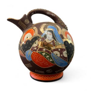 May include: A brown ceramic pitcher with a handle and spout, featuring a painted design of a woman in a blue headdress and gold accents. The pitcher has a round body and a decorative base with orange and red details.