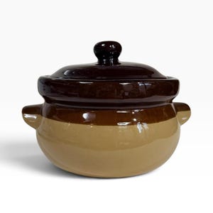 May include: A round ceramic bean pot with a glossy finish, featuring a two-tone design with a dark brown lid and upper rim, and a light tan base. The pot includes two small side handles and a matching lid with a central knob handle on top.