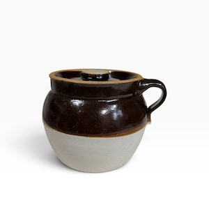 May include: A two-toned ceramic crock with a lid and a single side handle. The upper portion of the vessel features a glossy dark brown glaze, while the lower portion is finished in a matte, unglazed light beige or off-white clay texture.