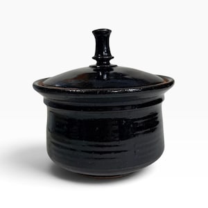 May include: A black glazed ceramic jar with a matching lid. The jar features a rounded body with subtle horizontal ridges and a wide rim. The lid has a central vertical handle with a flared top, creating a cohesive and dark monochromatic appearance.