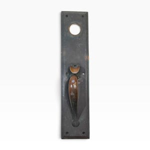 May include: A rectangular dark metal door plate featuring a circular opening at the top and a curved brown handle attached to the center. The plate has four small screw holes at the corners and shows signs of wear with visible surface scratches.