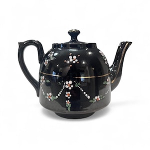 May include: Black ceramic teapot with a lid and handle. The teapot is decorated with white and orange floral patterns and gold trim. The spout and handle are curved. The teapot is a classic design.