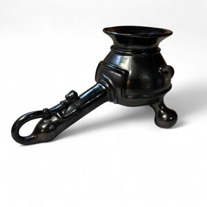 May include: A polished black ceramic vessel featuring a wide, flared rim, a rounded body with a small side handle, and three short, rounded legs. The handle is elongated and curved, ending in a loop, with decorative molded details along its length.
