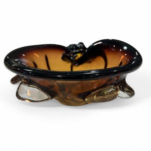 May include: A decorative glass ashtray with a unique design. The ashtray features a dark brown rim and a lighter amber-colored interior. It has a sculpted, organic shape with three clear glass feet and a textured detail in the center.