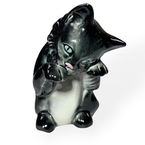 May include: A glossy black and white ceramic cat figurine. The cat is in a seated position, with its paws clasped together. It has blue eyes and a pink mouth. The figurine has a vintage aesthetic.