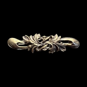 May include: A detailed, antique-style metal decorative element with an ornate, symmetrical design. The piece features stylized leaves and swirling patterns, with a slightly curved shape. The metal has a tarnished, aged appearance, suggesting it is vintage or vintage-inspired.