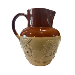 May include: A brown and tan ceramic pitcher with a handle and spout. The top and handle are a dark brown, while the body is tan with embossed windmill and village scenes. A classic piece for serving beverages.