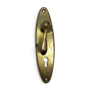 May include: An oval-shaped brass door handle plate with a weathered, scratched finish. The hardware features a central lever handle and a keyhole opening at the bottom, with two circular screw holes at the top and bottom for mounting purposes.
