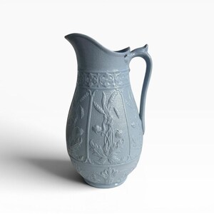 May include: A light blue ceramic pitcher featuring a textured surface with embossed floral and wheat motifs. The vessel has a curved handle, a wide pouring spout, and a decorative band around the neck, showcasing a consistent monochromatic finish.