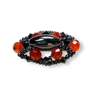 May include: An oval hair clip featuring a central smooth black stone surrounded by a decorative arrangement of small black beads, faceted black crystals, and vibrant orange translucent beads in a symmetrical, layered, and intricate design.