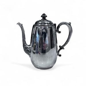 May include: A silver-colored coffee pot with a curved spout and ornate handle. The pot has a lid with a decorative finial. The surface of the pot reflects the surrounding environment. The design is vintage.