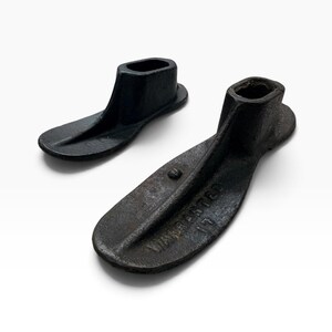 May include: Two black cast iron shoe lasts with a textured, weathered surface. The larger piece features the embossed text "WARRANTED" and the number "17" on its sole. These heavy metal tools have a distinct heel cup and a flat, elongated base.