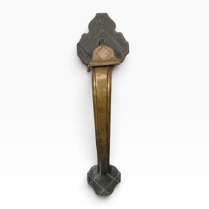 May include: A vertical brass door handle featuring a dark, weathered finish with a central gold-toned grip. The handle has decorative, clover-shaped mounting plates at the top and bottom, each engraved with a subtle grid pattern and a diamond shape.