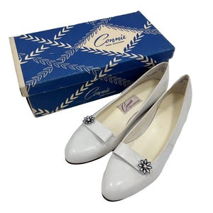 May include: A pair of white leather flats with a small flower embellishment, next to a blue shoe box. The box has a white leaf pattern and the brand name "Connie" in a decorative font. The shoes have a classic, vintage style.