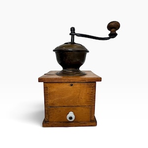 May include: A manual coffee grinder featuring a square wooden box base with a small pull-out drawer and a white ceramic knob. A dark metal hopper sits on top, equipped with a black metal crank handle that has a round wooden knob at the end.
