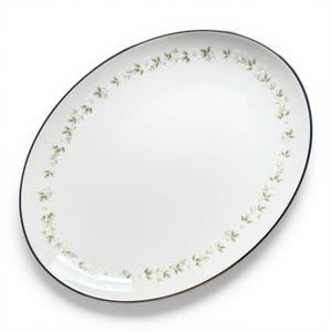 May include: An oval white serving platter featuring a delicate border pattern of small white flowers and soft green leaves. The rim of the platter is finished with a thin, dark-colored line that frames the floral design against the white surface.