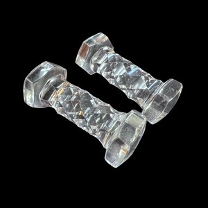 May include: Two clear, faceted glass dumbbells with hexagonal ends and diamond-patterned handles. The weights are displayed against a black background, highlighting their reflective surfaces. The design suggests a decorative or collectible item.