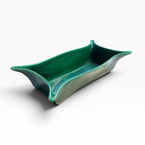 May include: A rectangular, emerald green ceramic dish with gently curved sides and flared, pointed ends. The dish has a glossy finish and a shallow interior, suitable for serving or display. The design is simple yet elegant.