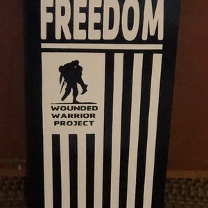 May include: A black and white wooden sign with the words "FREEDOM" and "WOUNDED WARRIOR PROJECT" and a silhouette of a soldier carrying another soldier. The sign has a design that resembles an American flag.