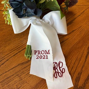 May include: White satin ribbon with a burgundy bow, embroidered with "PROM 2021" and a monogram. The ribbon is tied around a bouquet of dark blue and burgundy flowers.