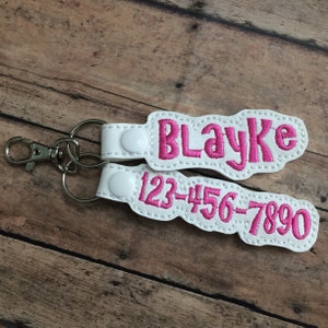 May include: A white faux leather keychain featuring two embroidered tags with pink stitching. The top tag displays the name Blayke, and the bottom tag shows the number 123-456-7890. Both tags are secured with white plastic snaps to a metal ring.