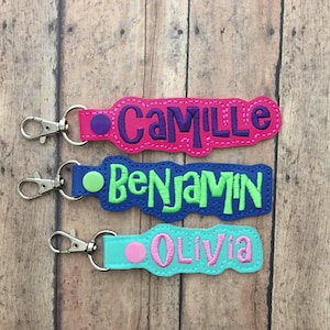 May include: Three personalized keychains with metal lobster clasps. The top is hot pink with purple embroidery reading Camille. The middle is blue with lime green embroidery reading Benjamin. The bottom is light blue with pink embroidery reading Olivia.