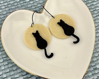 May include: A pair of cream-colored cat silhouette earrings with black cat cutouts. The earrings are on a white heart-shaped plate.