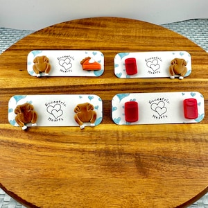 May include: Four sets of Thanksgiving-themed earrings on a wooden surface. Each set features a white card with blue hearts and the text "Eccentric Hearts." The earrings include turkey, pie, and red can designs.