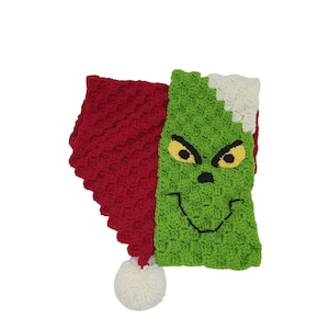 May include: A crocheted Grinch-themed blanket with a red and green color scheme. The Grinch's face is embroidered on the green side of the blanket. The blanket has a white pom-pom on the end.