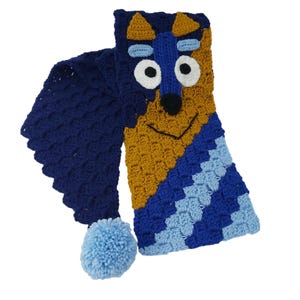 Op de afbeelding: Een handgemaakte, blauwe, gele en witte gehaakte sjaal met een hondengezichtontwerp. De sjaal heeft een pompon aan het uiteinde en een gestructureerd patroon. Het honden gezicht heeft grote witte ogen, een zwarte neus en een lachende mond.